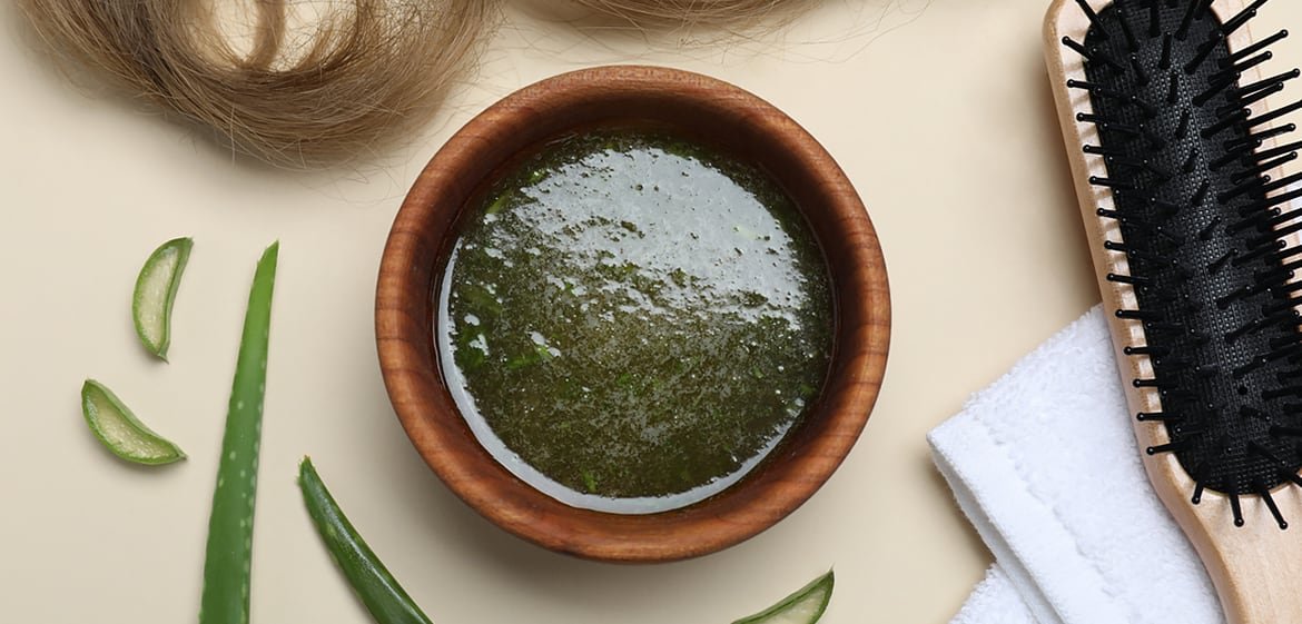 Braune Schale mit grünem Haaröl, ein Handtuch, Aloe-Vera, eine Haarbürste und ein brauner Haarschopf auf einem Tisch